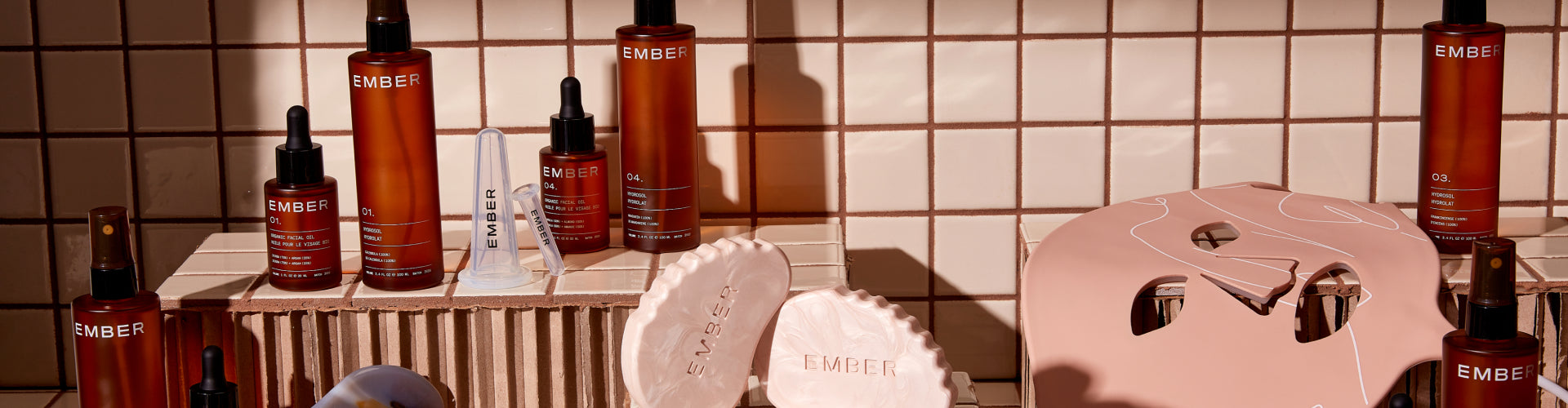 A collection of Ember products arranged on tile risers in front of a tiled background. Some products include oils, hydrosols, the LED Light Therapy Mask, cupping set and more.