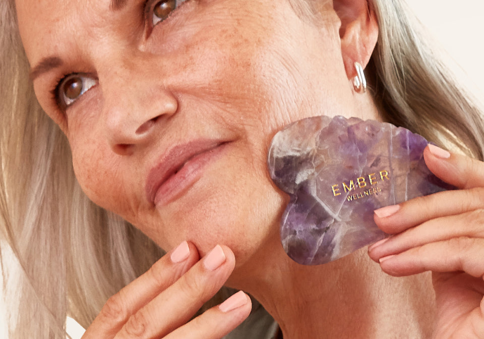 Our patented Heart Gua Sha tool designed for optimal facial support, seen here in amethyst being used by a woman with light hair.