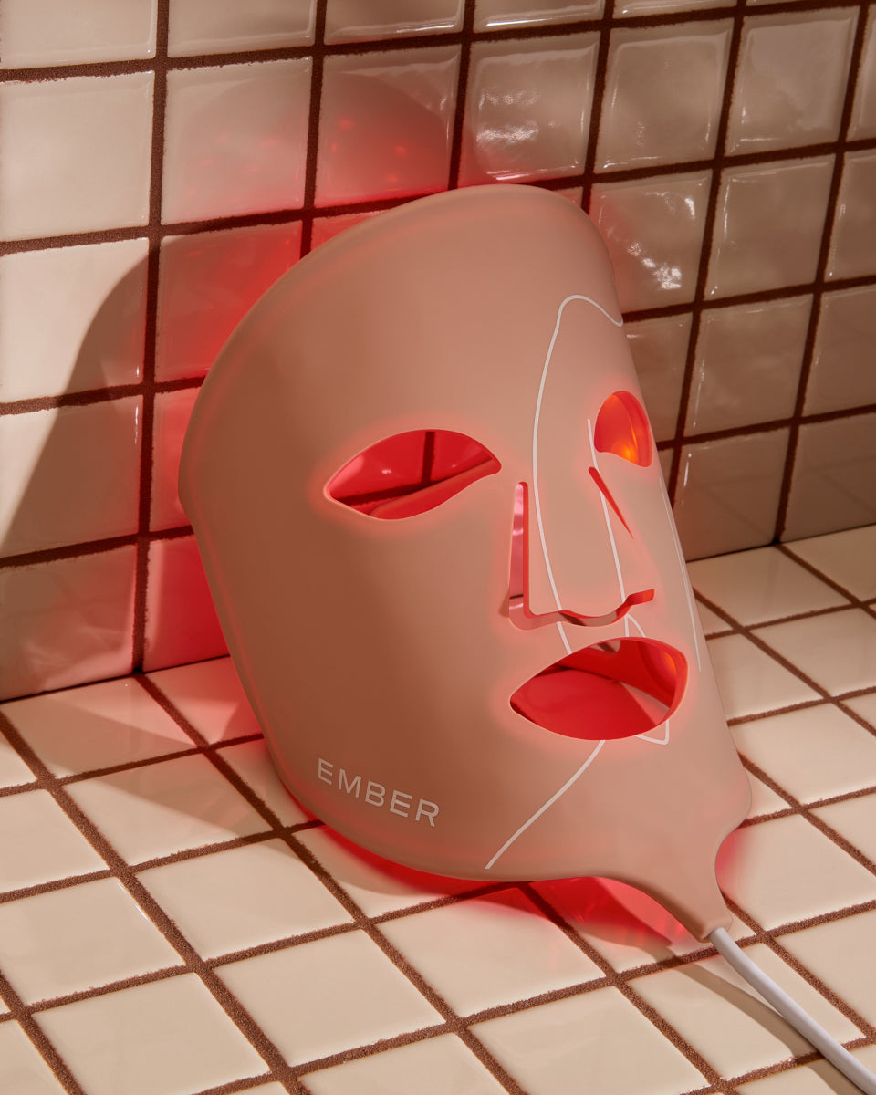The Ember LED Light Therapy Mask, emanating a red glow, rests on a tiled surface.