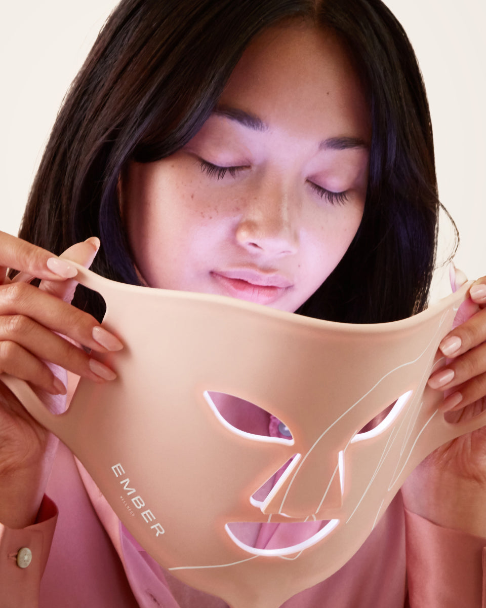 A woman in a pink top holds the Ember Wellness LED Light Therapy Mask to her face.