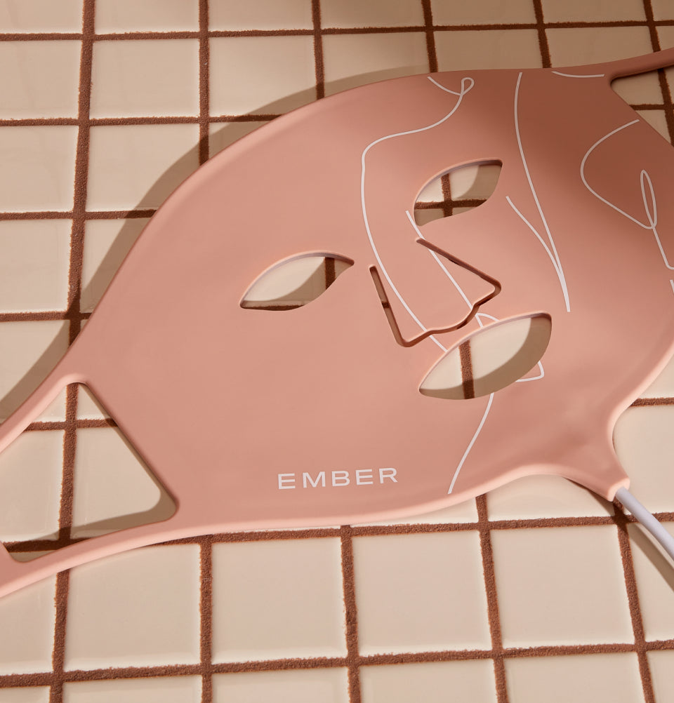 Ember's LED Light Therapy Mask in pink rests on a tiled surface. This patented, easy-to-wear mask is made of silicone and easily wraps around your face with an adjustable velcro strap. 