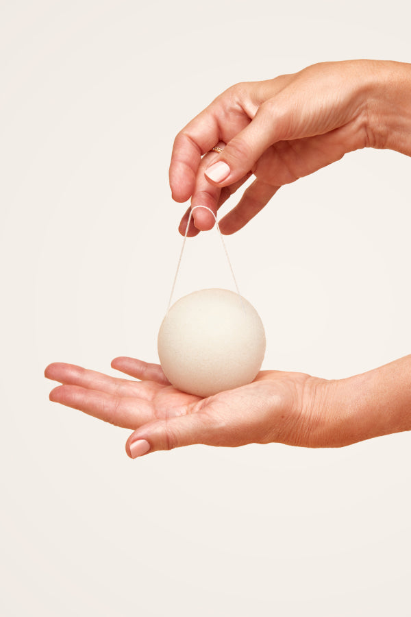 Embers Konjac Sponge, an eco-friendly, all-natural, gentle, and reusable facial cleansing sponge made from vegetable fibre. Shown held in the palm of a women's hand, suspended by her other hand on its attached string.